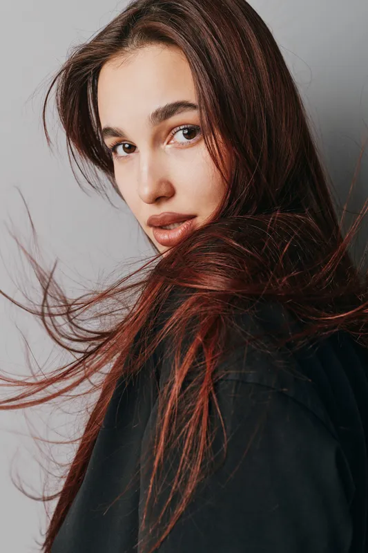 Young woman with long brown hair looking over shoulder, wearing black clothing against plain light background. Portrait, beauty, natural makeup, casual style, confident expression.