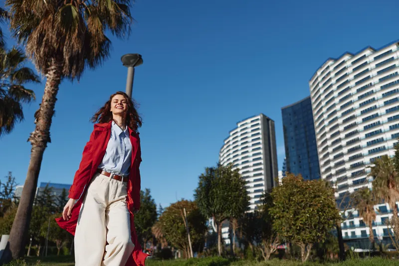 A young woman with curly hair in a flowing red coat walks through a sunny city park, exuding confidence and style against palm trees and modern buildings.