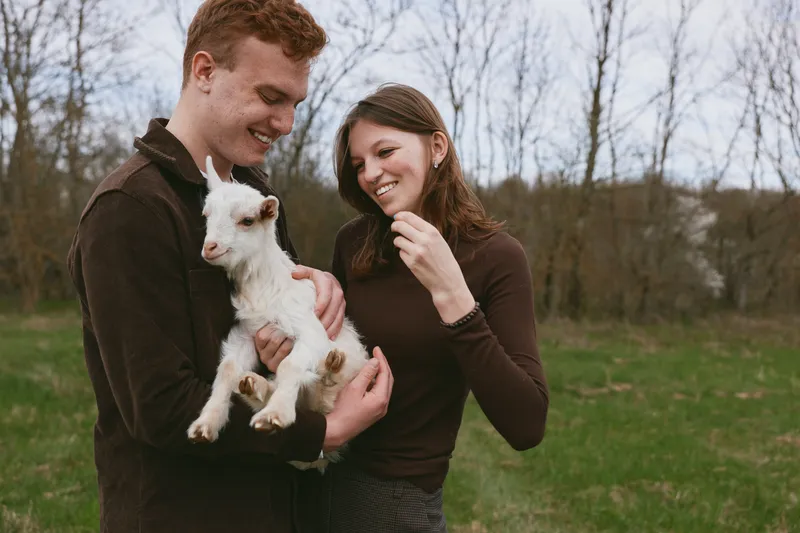 Cheerful couple outdoors, smiling while holding a goat kid, warm sunlight, casual outfits, pastoral setting with trees and grass, fresh air, candid lifestyle moment, natural colors, connection