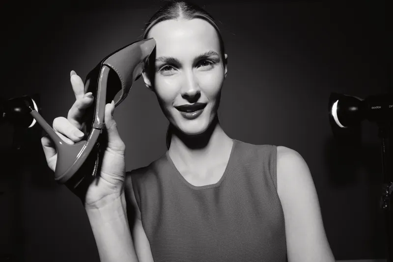 Elegant white woman with light skin holds high heel shoe near face smiling in black and white studio portrait. Concept of fashion, style, and footwear.