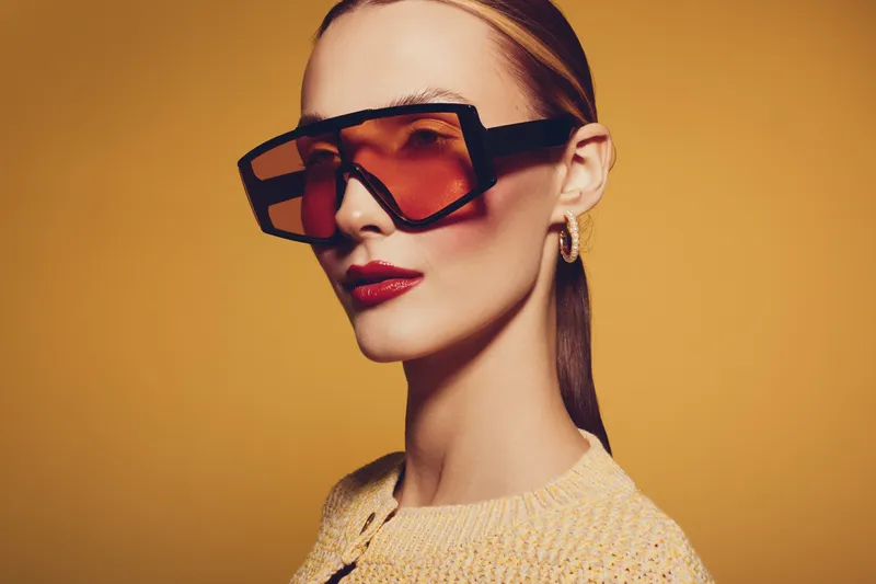 Stylish woman with slicked back hair wearing oversized tinted sunglasses and red lipstick poses against yellow background. Concept of fashion, beauty, accessories, and modern style.