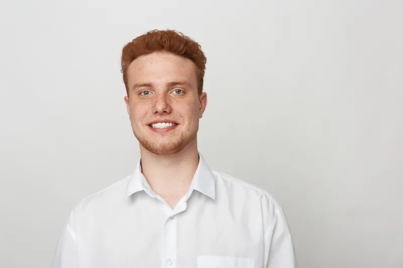 Smiling young man with red hair and freckles wears white shirt, posing against plain light background. Portrait of confident male model with casual style and friendly expression.