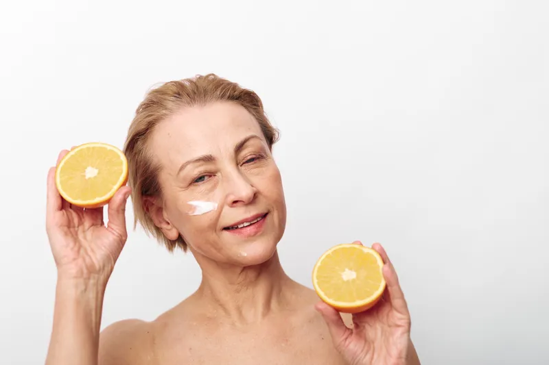 Smiling mature woman holds two orange halves near face with white cream on cheek. Bright studio background highlights natural skincare, healthy lifestyle, and citrus fruit benefits concept.