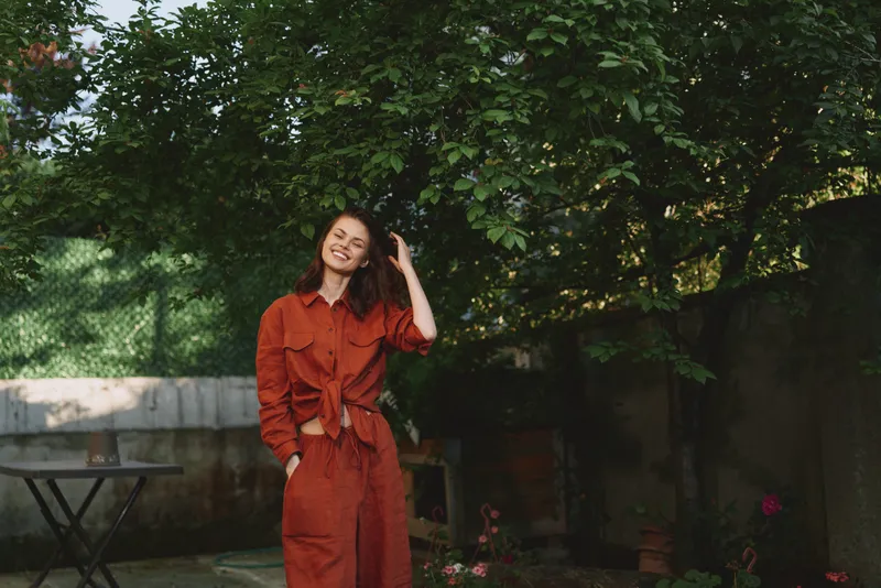 Cheerful woman outdoors in a warm orange outfit, smiling softly, relaxed pose, sunlit garden scene, lush greenery, natural colors, candid lifestyle moment, carefree mood