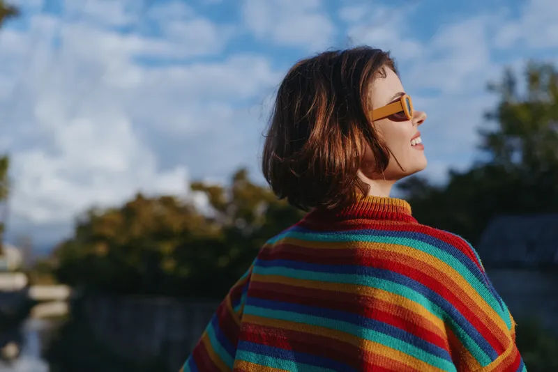 Woman in her late twenties wearing a colorful striped sweater and yellow sunglasses smiling and looking up outdoors by riverbank, short brown hair, relaxed travel lifestyle, sunny autumn mood