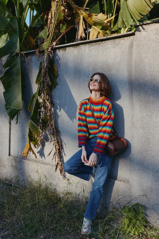 woman striped sweater sunglasses leaning against a tropical wall, smiling and relaxed. Young adult with short hair in blue jeans and crossbody bag, casual outfit for travel lifestyle.