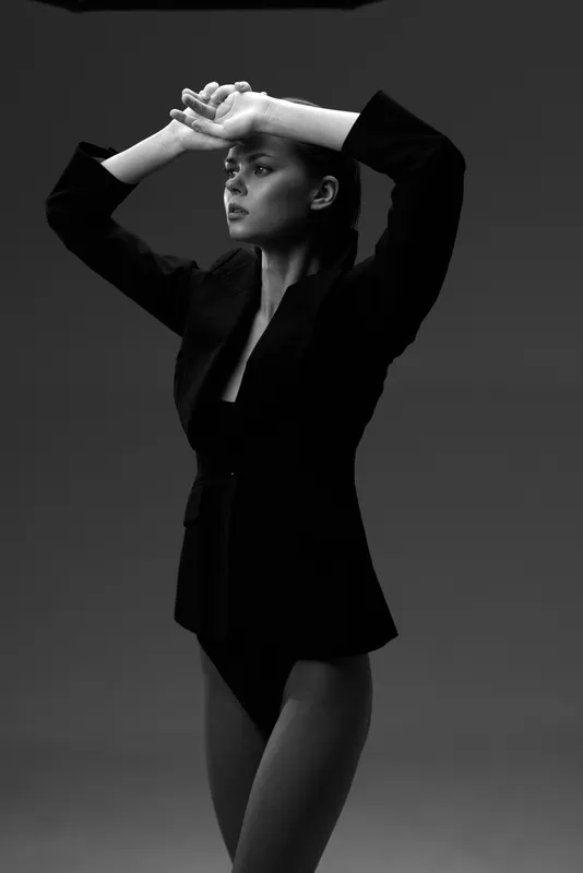 Black and white portrait of a stylish woman posing with raised arms wearing a blazer and bodysuit.