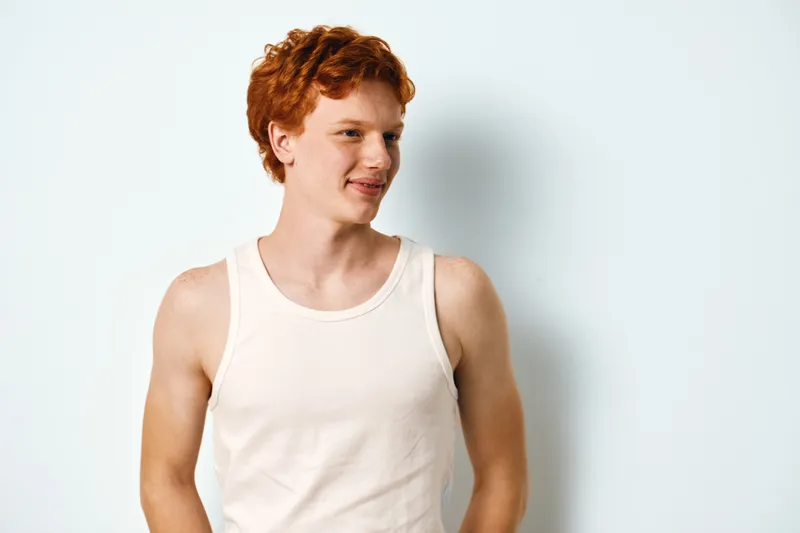 Young man with red hair wearing a white sleeveless shirt standing and smiling against plain white background looking aside for casual portrait or lifestyle concept.