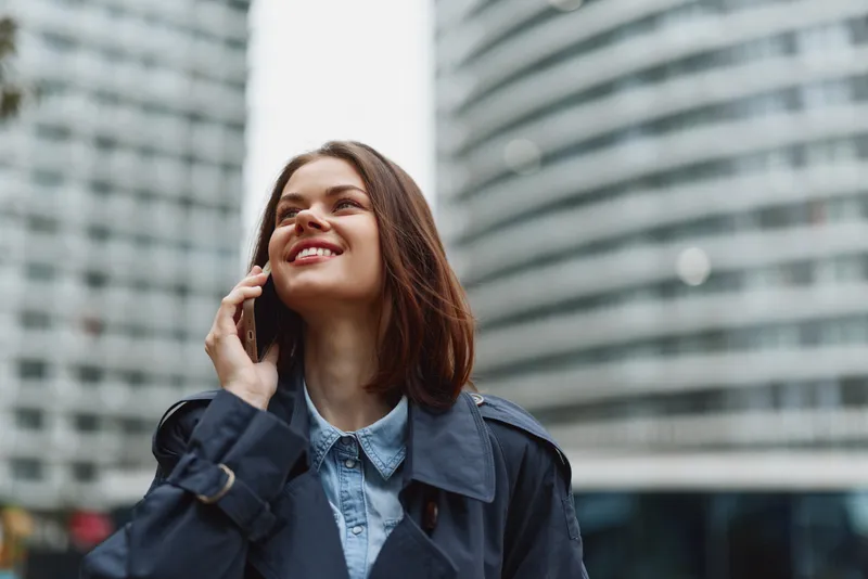 Young woman in blue coat talks on smartphone outdoors in city, smiling and looking up with modern office buildings in background, casual urban lifestyle concept.