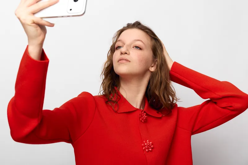 Young woman in red sweater takes a selfie with smartphone, posing confidently against white background. Concept of social media, self-expression, and modern lifestyle photography.
