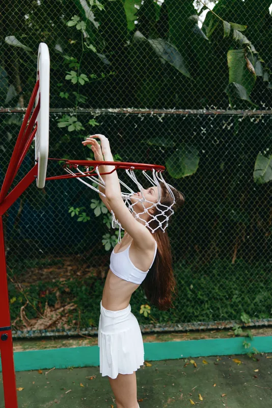 Young woman in sporty attire joyfully reaching for a basketball hoop, showcasing athleticism and energy in a vibrant green outdoor setting