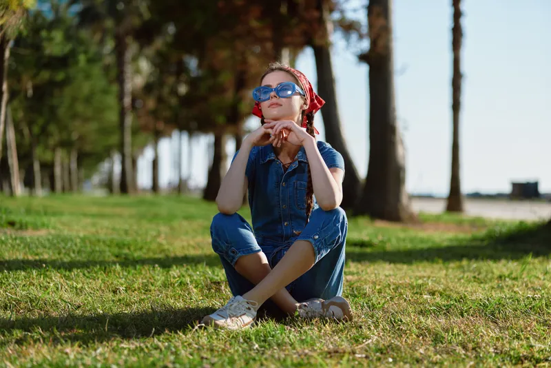 Sitting young woman with sunglasses and red scarf enjoying sunny day in green park with trees, casual outfit, relaxed and peaceful mood outdoors.