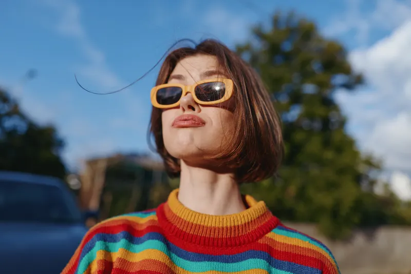 woman in striped sweater with yellow sunglasses and short brown hair shows a playful expression outdoors on a sunny day in a suburban street, casual outfit, confident pose for lifestyle travel imagery