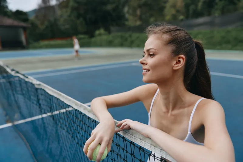 Young athletic woman smiling at the tennis court, wearing a white sports outfit, surrounded by greenery and blue courts, representing a healthy lifestyle and outdoor sports