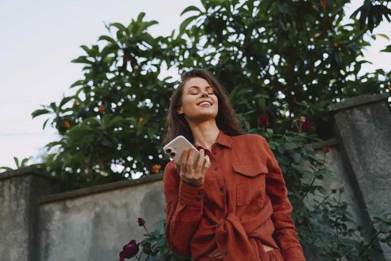 Woman lifestyle shot outdoors wearing an orange dress, smiling while holding a smartphone, standing near greenery and a wall, candid cheerful mood, relaxed confident vibe in soft sunlight, modern