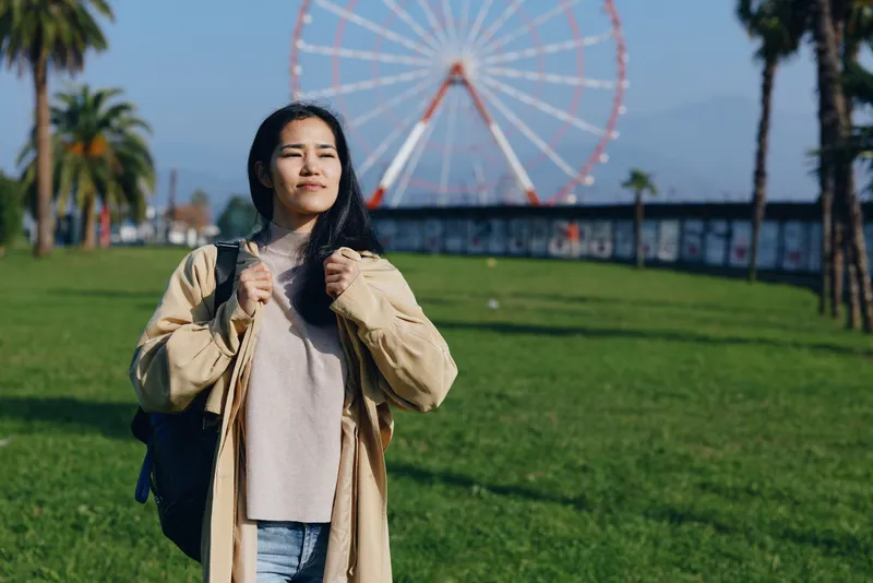 Woman in a casual jacket stands in a sunny park with a ferris wheel in the background, backpack ready for travel, outdoors, relaxed mood, and a candid urban leisure scene