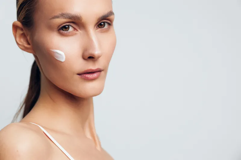 Young woman with clear skin applies moisturizing cream on her cheek while looking calmly into the camera against a plain light background.