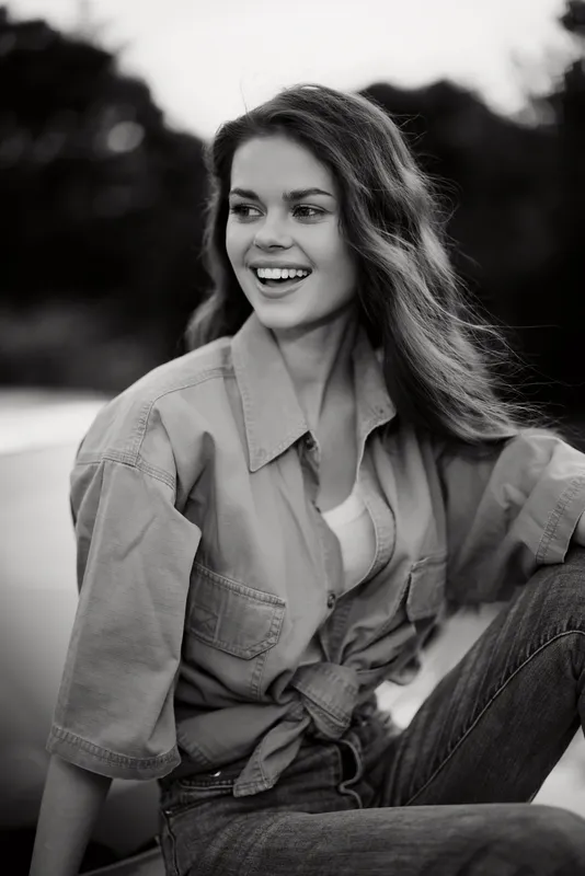 Young woman sitting outdoors in a casual outfit smiling, exuding happiness and confidence, surrounded by nature in black and white.