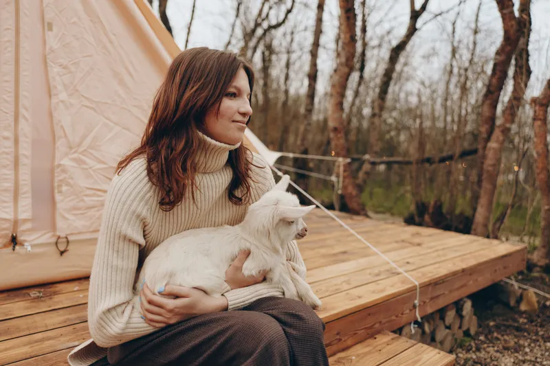 Caring woman holding a goat kid beside a tent in a woodland campsite. Cozy sweater, warm light, lifestyle scene focused on animal bonding, outdoor calm, nature welcome, gentle affection.