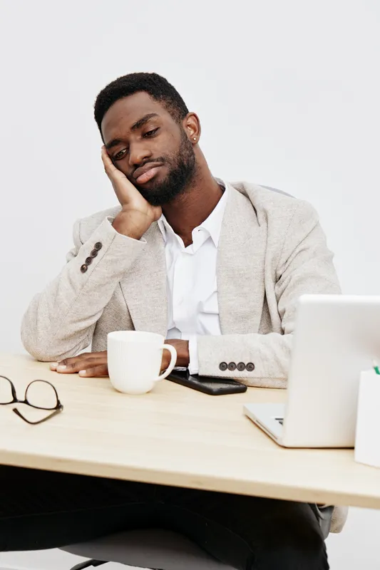 Thoughtful young man with tired expression sitting at desk with coffee mug and laptop, resting his head on hand in office workspace, wearing smart casual outfit, indoor shot, concept of fatigue or str