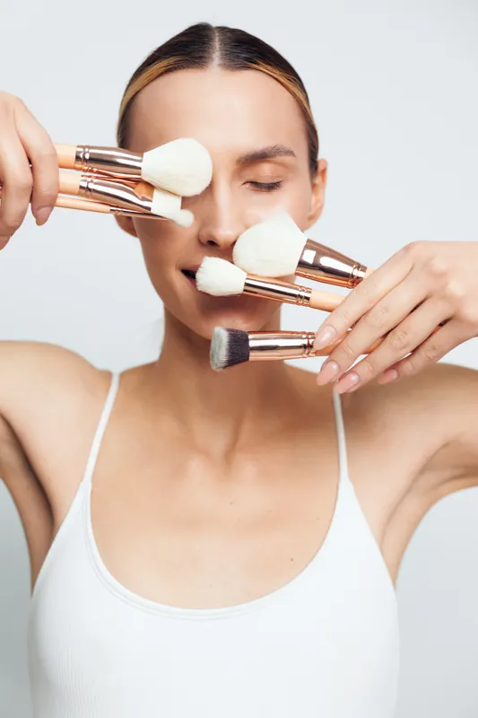 A young woman in a white tank top holds multiple makeup brushes close to her face with eyes closed. The image highlights beauty tools and skincare routine concepts.