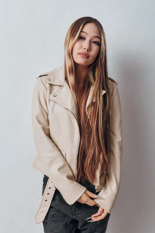 Young woman with long straight hair wearing beige leather jacket and black pants posing with calm expression on white background for fashion and style concept.
