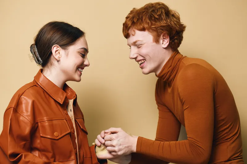 Young couple smiling and holding hands together with joy and connection, expressing happiness and love in casual warm clothing at studio background.