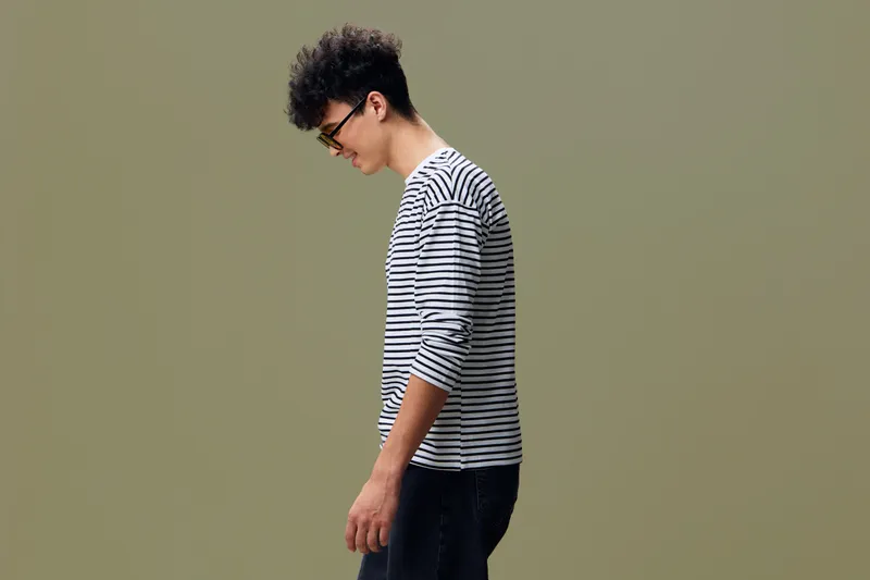 Young man in striped shirt standing sideways with downward gaze, casual style, curly hair, neutral background, thoughtful mood, minimalist setting.
