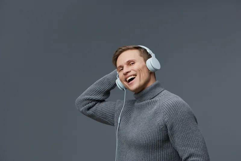 Smiling young man wearing gray sweater listening to headphones, looking happy and relaxed against plain gray background, studio portrait, lifestyle concept, casual style, positive mood, enjoying music