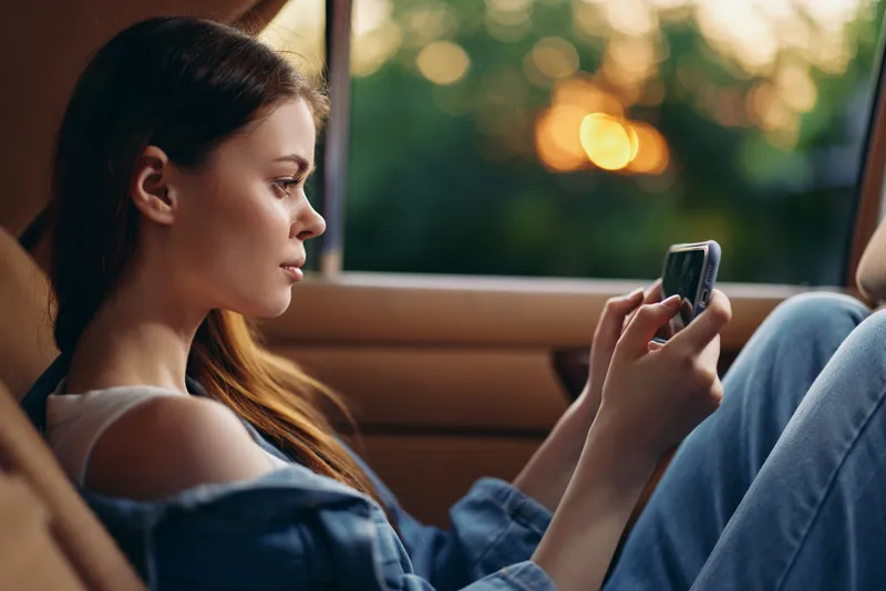 Young woman using her smartphone in a car during sunset, capturing a serene moment with relaxed emotions and casual attire.
