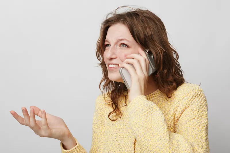 Young woman with curly hair wearing yellow sweater talks on smartphone with expressive gesture. Isolated on white background. Concept of communication, technology, and casual lifestyle.