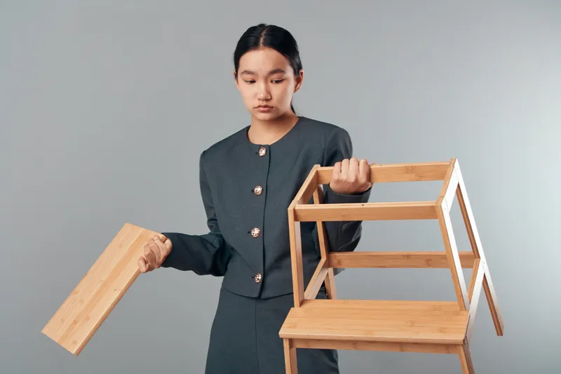 Young woman in formal clothing holding a stackable wooden shelf with a panel removed, standing against a neutral background for interior design or furniture concept.