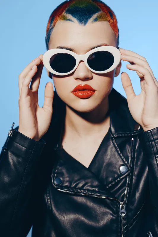 Stylish woman with short colorful hair wearing white sunglasses and black leather jacket, posing confidently against a dark background with bold makeup.