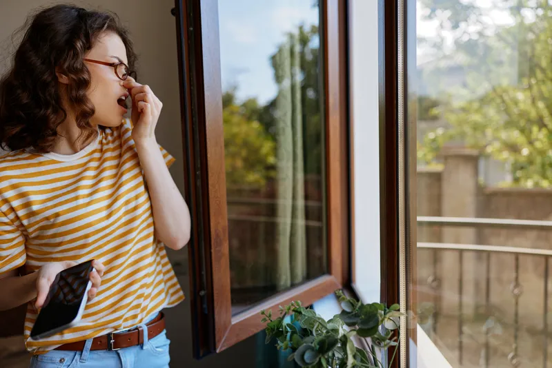 Candid woman indoors by a window, lifestyle moment at home, striped shirt, natural light, greenery outside, relaxed pose, warm colors, cozy atmosphere