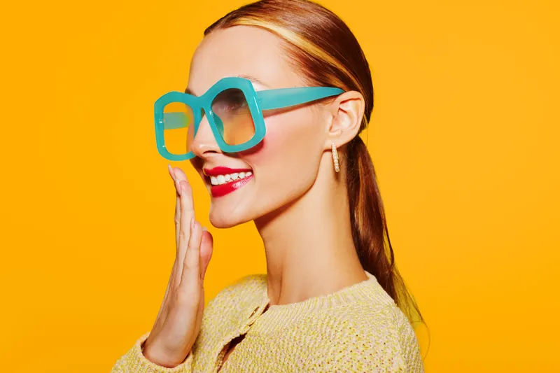 Smiling woman with red lipstick and large turquoise sunglasses poses on bright yellow background. Fashion portrait with colorful accessories and trendy style concept.