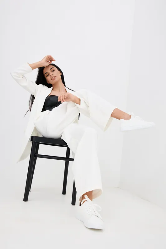 relaxed woman in white suit sitting on black chair in minimalistic white studio, wearing black top and white sneakers with casual confident pose