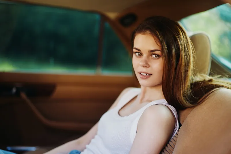 A young woman with long hair sitting in a vintage car, wearing a casual white tank top, smiling confidently against a lush green background.