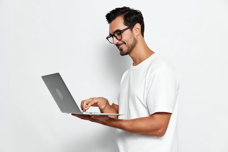 Young smiling man using laptop, wearing casual white t-shirt, looking focused, isolated on plain light background. Modern technology lifestyle concept.