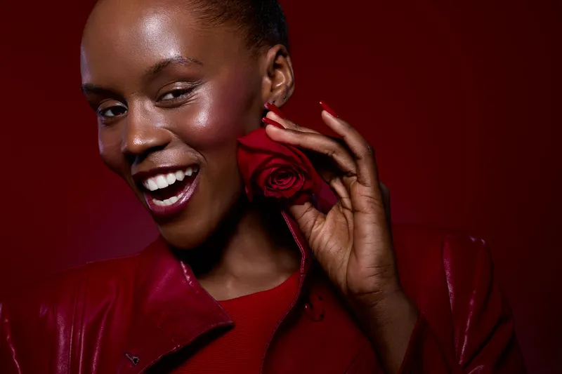 Close-up of happy black woman with dark skin holding red rose near face smiling confidently on red background. Concept of beauty, elegance, and natural skin glow in studio lighting.