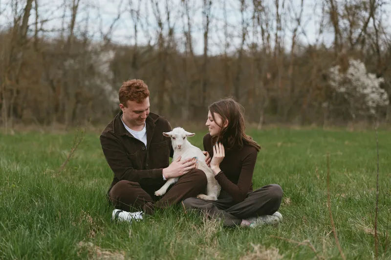 A loving couple sits in a green meadow, cradling a playful goat kid between them. An outdoor lifestyle moment in bright natural light, amid open field and rural nature.