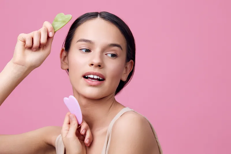 Young woman with gua sha tools for facial massage, showcasing healthy skincare on a pink background, expressing joy with a radiant smile