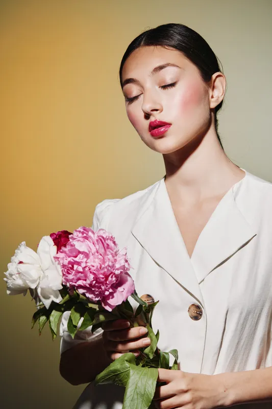 Young woman with bright red lipstick holds white and pink peony flowers in beige studio. Styled in minimal white dress, eyes closed, embodying fashion beauty creativity concept.