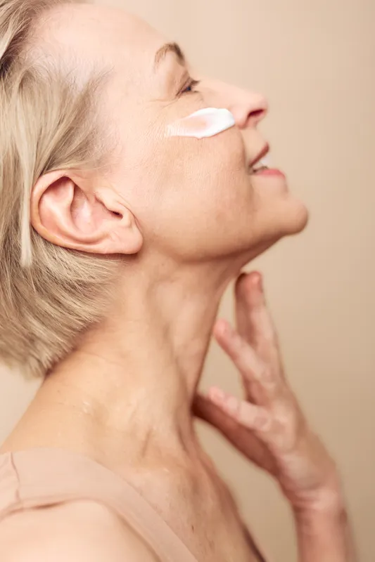 Mature woman with short blonde hair applies face cream on cheek, smiling gently. Close-up on beige background. Concept of skincare, aging, beauty, and self-care routine.