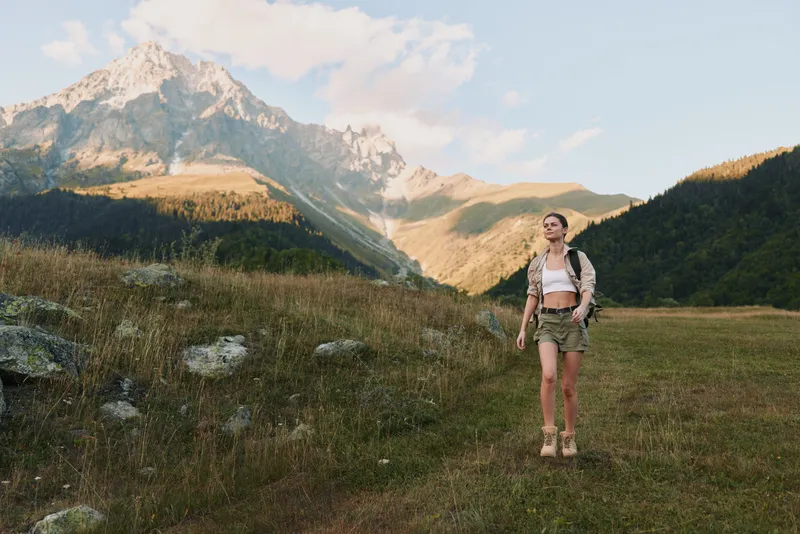 Young woman hiking in mountains wearing shorts and backpack walking on grassy trail under clear sky with rocky peaks in background enjoying active lifestyle outdoors.