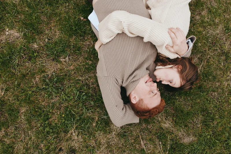 A couple lying on grass in a park, embracing softly, smiling, enjoying a quiet outdoor moment, casual sweaters, tender connection, candid lifestyle scene, peaceful and intimate.
