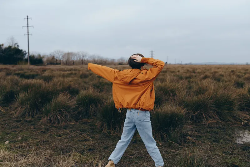 Woman walking outdoors through grassy field, orange jacket and blue jeans, hood up against cloudy sky, relaxed casual lifestyle scene, soft overcast light, calm nature mood