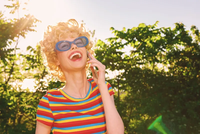 Vibrant woman outdoors in a striped shirt and retro sunglasses, laughing in bright sunlight, joyful mood, casual style, nature behind, capturing a carefree lifestyle moment