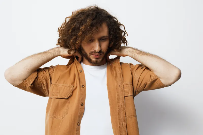 Young man with curly hair in a brown shirt expressing contemplation, set against a minimalistic white background, conveying a modern lifestyle concept
