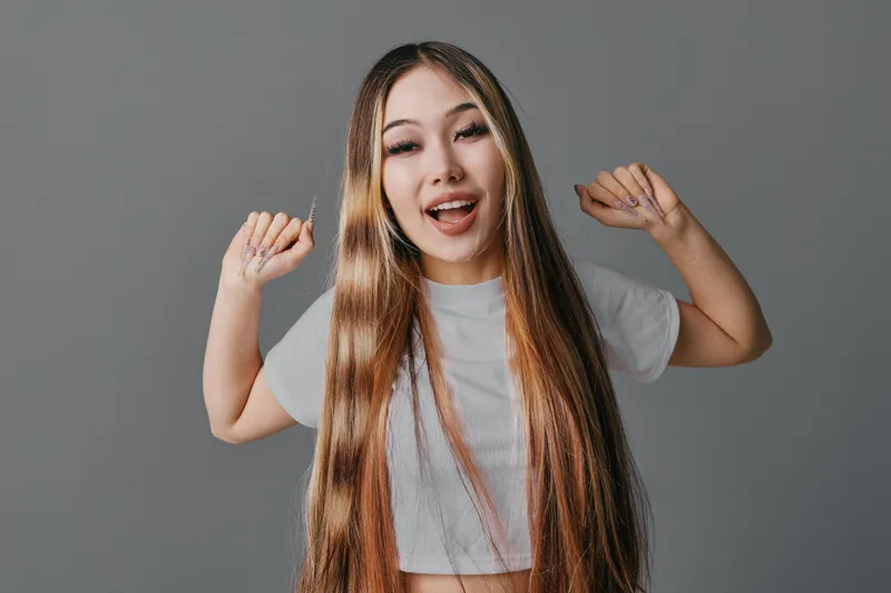 Young woman with long hair smiling and posing with raised hands on gray background. Female model expressing happiness and confidence in casual white t shirt indoors.
