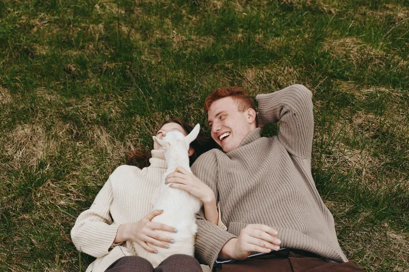 A relaxed couple lies on grass with a curious goat kid nearby, enjoying carefree outdoors. A lifestyle moment capturing bonding, playfulness, and nature in a cozy countryside setting.